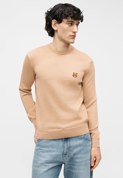 Jeune homme aux cheveux bouclés et foncés portant un pull beige avec un petit logo de renard, associé à un jean bleu clair, debout devant un fond uni.