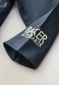 Tumed sinine puuvillane varrukas ribakujundusega ja heledas beežis tikitud logo "BAKER". Omab ümarat mansetti koos õmblusteadetega.