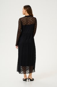 Rochie din dantelă neagră cu mâneci lungi, spate transparent și fustă fluidă. Prezintă un model floral complicat și un underskirt până la genunchi.