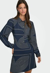 Marineblauwe jurk met lange mouwen, kraaghalslijn en knoopsluiting. Heeft geometrische witte lijnpatronen door het geheel. Glad stoftextuur.