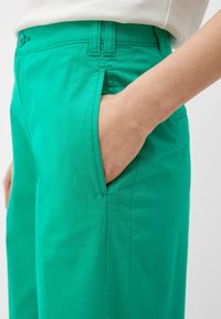 Grüne Hose aus leichtem Material mit glatter Textur, Hosentaschen und genähten Nähten. Der Bund ist mit Gürtelschlaufen ausgestattet.