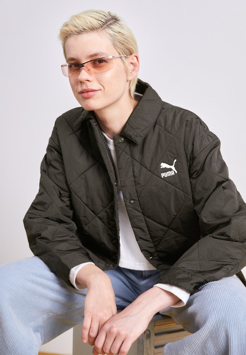Svart quilted jakke med krage og hvit Puma-logo, kombinert med en hvit skjorte og lyseblå cordfløyelsbukser. Glatt stofftekstur.