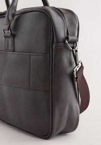 Braune Lederaktentasche mit strukturierter Oberfläche, obenliegendem Griff, abnehmbarem Riemen und silberner Hardware sowie einer Taschenverzierung auf der Vorderseite.