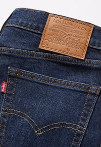 Mörkblå denimjeans med en tan läderlapp med präglad logotyp, gula sömmar och en röd Levi's-etikett på bakfickan.