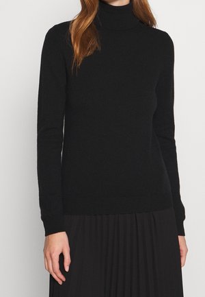 Frau trägt einen enganliegenden schwarzen Rollkragenpullover mit langen Ärmeln und einen schwarzen plissierten Rock, stehend vor einem schlichten hellen Hintergrund.