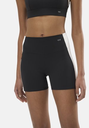 Mujer con pantalones cortos deportivos negros de cintura alta con paneles laterales blancos y un sujetador deportivo negro a juego, de pie frente a un fondo blanco.