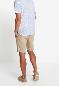Camicia a maniche corte a scacchi blu chiaro abbinata a pantaloni beige, che mostra un taglio rilassato e uno stile casual. Scarpe marroni con lacci visibili.