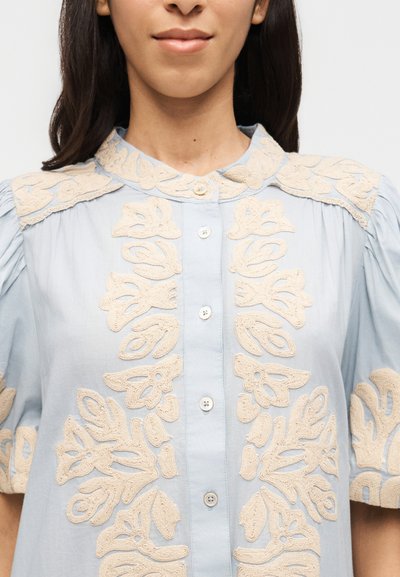 Γυναίκα που φοράει ένα ανοιχτού μπλε πουκάμισο με κουμπιά, διακοσμημένο με μπεζ δαντελωτά λουλουδένια κεντήματα στο γιακά, μπροστά, στα μανίκια και στους ώμους.