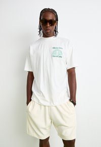 T-shirt en coton blanc avec un texte graphique vert sur le devant, assortie à un short crème clair. Modèle portant des lunettes de soleil marron.