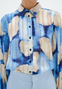 Blouse à col boutonnée avec un motif abstrait bleu, beige et blanc, associée à un pantalon taille haute bleu clair.