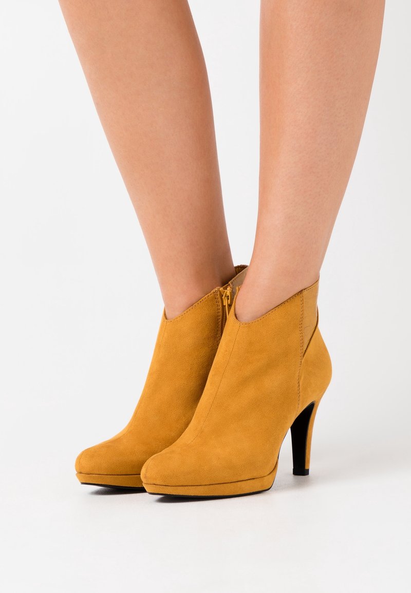 Femme portant des bottines en daim jaune moutarde avec une fermeture éclair latérale et des talons aiguilles hauts, debout sur une surface blanche.