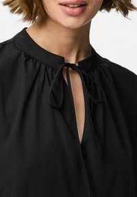 Mustamet särk ümmarguse kaelusega, millel on võtmeaugu avamine ja lipsu detail kaelas. Pehme kangas, millel on kerge kogumine õlgadel.