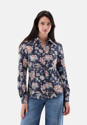 Donna con i capelli lunghi e scuri che indossa una camicia a maniche lunghe blu navy a fiori con bottoni e jeans blu, in piedi davanti a uno sfondo uniforme.