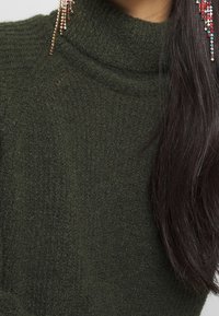 Långt mörkt hår över en texturerad mörkgrön polotröja, med flerfärgade pärlörhängen som hänger synligt på ena sidan.