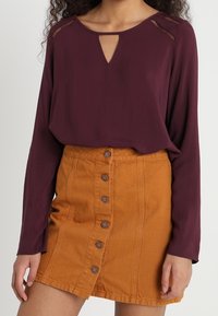 Blusa bordeaux a maniche lunghe con dettaglio a occhiello abbinata a una gonna gialla senape con bottoni, realizzata in denim con tessuto testurizzato.