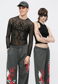 Schwarzes, durchsichtiges Langarmshirt mit komplizierten Mustern, graue Jogginghosen mit rotem Grafikdesign und ein schwarzes Tanktop; beide tragen lässige Mützen.