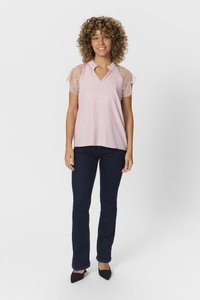 Blusa rosa chiaro con maniche corte in pizzo, scollo a V, abbinata a jeans blu scuro e tacchi a punta neri. Tessuto liscio, vestibilità comoda.