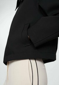 Zwarte jas met een cropped pasvorm, voorzien van een gladde textuur en zijzakken. Gecombineerd met lichtbeige broeken met zwarte biesaccenten.