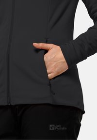 Zwarte softshell jas met rits, vlakke naden en een hand in een zijzak. De stof lijkt glad en flexibel. Logo zichtbaar.