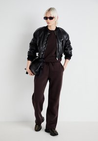 Black leather bomber jacket, brown sweatshirt, matching jogger pants, black bag with tie detail, and patterned black shoes.