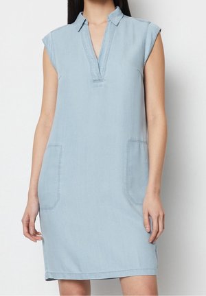 Bezrękawna, jasnoniebieska sukienka dżinsowa z kołnierzykiem, dekoltem w serek i przednimi naszywanymi kieszeniami, noszona przez stojącą osobę.