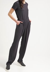 Musta haalari, jossa on lyhyet hihat, joustava vyötärö ja sivutaskut. Valmistettu sileästä kankaasta, jossa on rento istuvuus ja suppilomaiset lahkeet.