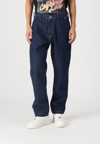 Jeans de mezclilla azul oscuro con un ajuste holgado, que presentan costuras anaranjadas en contraste, cinco bolsillos y un cierre de botón. Llevados con zapatillas blancas.