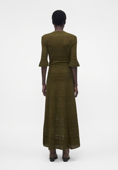 Άτομο με κοντά σγουρά μαλλιά, φοράει ένα ελαιί πράσινο, πλεκτό φόρεμα με μοτίβο και 3/4 μανίκια και καφέ μποτάκια μέχρι τον αστράγαλο, στραμμένο με την πλάτη.