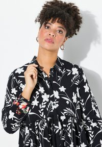 Abito nero con bottoni, decorato con un motivo floreale bianco, vita arricciata e accenti floreali colorati sulle maniche. Tessuto dalla texture morbida.