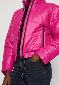 Chaqueta corta de plumón fucsia con acabado brillante, con bolsillos con cremallera y cuello alto. Fabricada con material ligero e aislante.