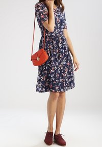 Marineblaues Blumenkleid mit kurzen Ärmeln und tailliertem Schnitt, kombiniert mit einer kleinen roten Umhängetasche und bordeauxroten Schuhen.