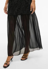 Juodas maksi sijonas su tekstūruotu viršutiniu sluoksniu, skaidriu plazdančiu apatine dalimi ir paprastais juodais sandalais su juostelėmis, turinčiais auksinius akcentus.