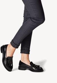 Sorte patentlæder loafers med kvastdetaljer, rund tå og lav stablet hæl, parret med mørke tætsiddende bukser, der er rullet op i kanten.