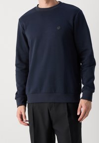 Námořnická modrá mikina vyrobená z hladké tkaniny, s kulatým výstřihem, žebrovanými manžetami a malým emblémem na hrudi.