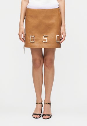 Femme portant une mini-jupe en cuir beige avec des lettres en corde blanches formant "OSC" et des sandales noires à talons hauts avec des boucles dorées.
