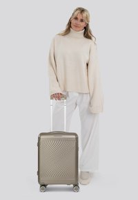 Harde koffer in metallic champagne, met een golvende textuur, witte handgreep en vier draaiwielen, vastgehouden door een persoon in lichte kleding.