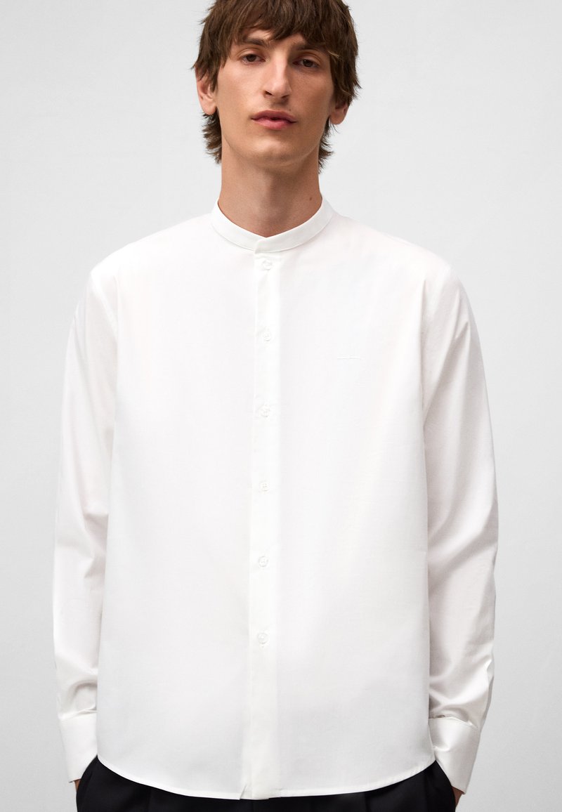 Vit långärmad skjorta med bandkrage, framsidan med knappstängning och en slät yta. Har en minimalistisk design utan mönster.