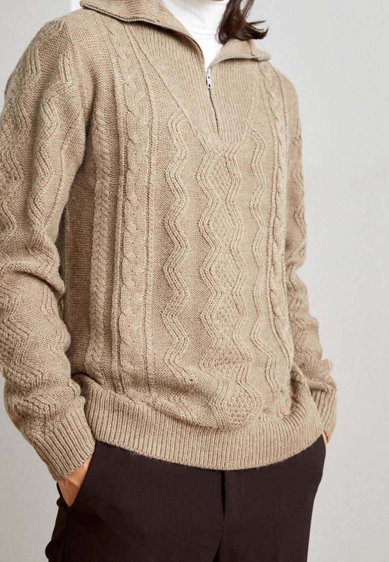 Beiger gestrickter Pullover mit einem Reißverschluss am Kragen, der strukturierte Zopf- und Zickzackmuster aufweist. Gerippte Bündchen und Saum, kombiniert mit einer dunklen Hose.