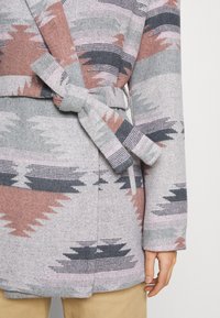 Abrigo de tejido en gris, rosa y marino con patrones geométricos. Incluye un cinturón atado y una tela suave y texturizada.