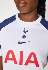Bijela Nike majica ima mornarsko plave akcentne detalje, logo Tottenham Hotspura i veliki crveni natpis "AIA". Tkanina ima prozračnu teksturu.