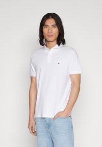 Polo bianco in cotone, con colletto a due bottoni e un piccolo logo sul lato sinistro del petto. Abbinato a jeans azzurri chiari.