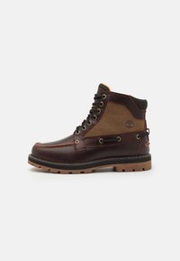 Brown leather boot with a tan fabric panel, round toe, metal eyelets, black rubber sole, and stitching details. Features raised logo on side.