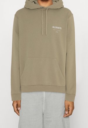 Osoba ubrana w jasnobrązową bluzę z kapturem, kieszenią z przodu i logo „ALLSAINTS” na lewej górnej części klatki piersiowej, do tego jasnoszare spodnie.