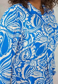 Blue and white floral patterned dress with a collar, buttons, and long sleeves; smooth fabric with visible intricate designs.