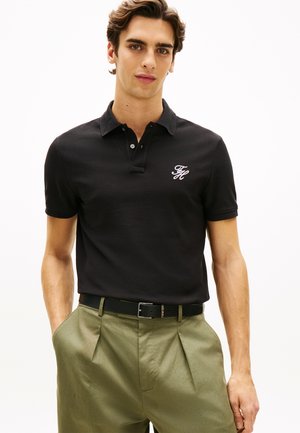 Νέος άνδρας που φοράει μαύρο πόλο μπλουζάκι με λευκό λογότυπο και ελαιδί πλισέ παντελόνι, με τα χέρια στις τσέπες, στέκεται μπροστά σε λευκό φόντο.