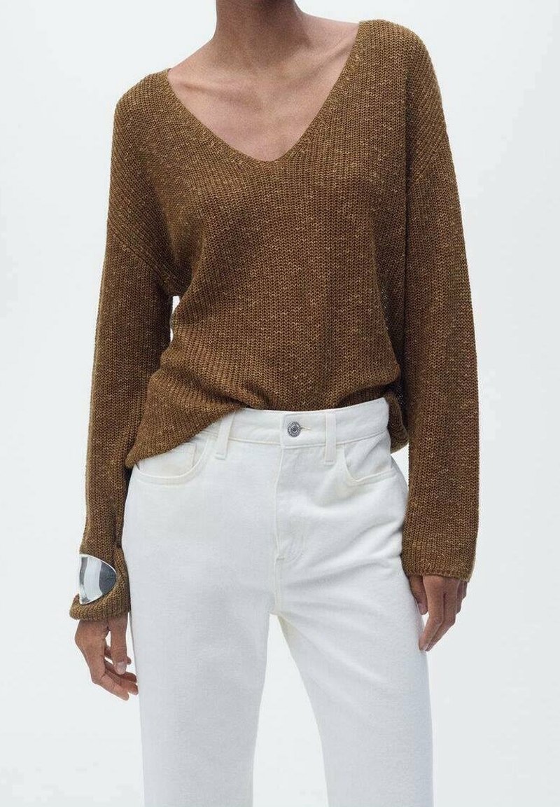 Femme portant un pull marron à col en V en maille avec des trous pour les pouces et un pantalon blanc taille haute, sur un fond uni.