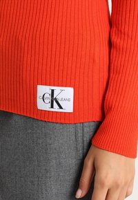 Pull orange côtelé avec une étiquette blanche portant l'inscription "CALVIN KLEIN JEANS" et un logo noir, porté sur un tissu gris, mettant en valeur les contrastes de couleurs.