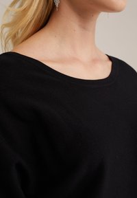 Černý, žebrovaný pletený top se širokým výstřihem a jemnou texturou. Detailní záběr oblasti ramen a klíční kosti, minimalistický design.