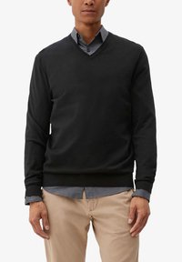 Schwarzer V-Ausschnitt-Pullover aus einem weichen, strukturierten Stoff. Getragen über einem hellgrauen Hemd mit Knopfleiste, mit sichtbaren Manschetten und einer lockeren Passform.