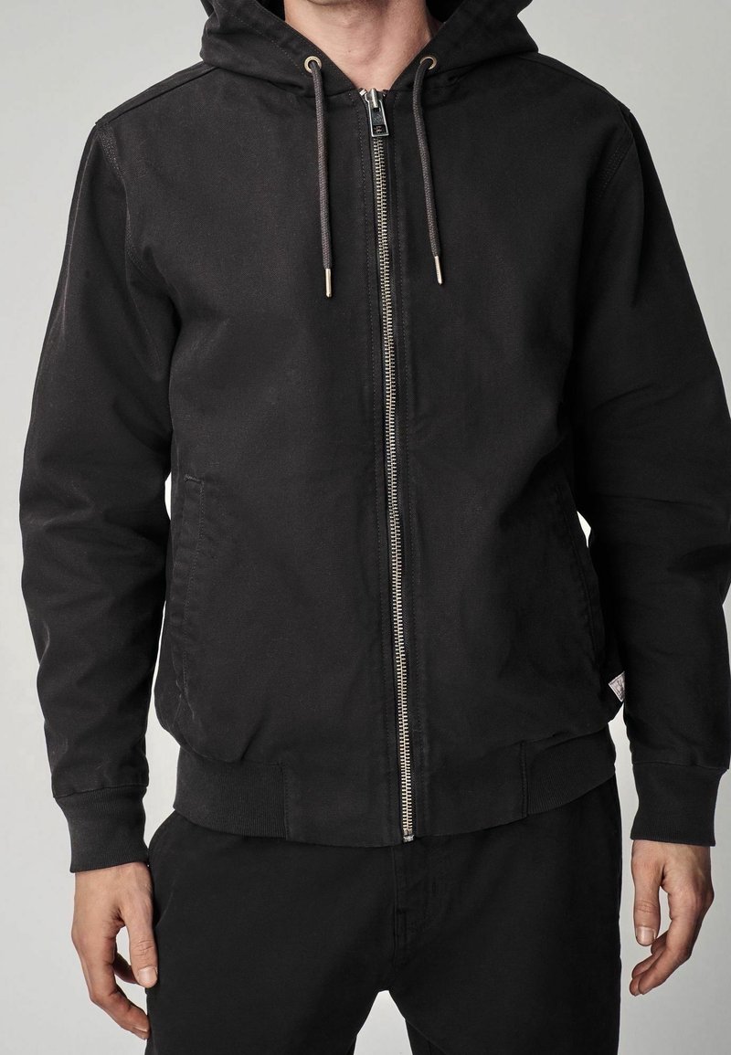 Homme portant un sweat à capuche noir avec fermeture éclair et cordons, debout les bras détendus le long du corps devant un fond uni.